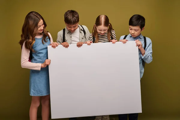 Positive multiethnische Preteen-Kinder mit Rucksäcken lächeln, während sie zusammen stehen und leere Plakate auf khakifarbenem Hintergrund betrachten, Happy Children 's Day Konzept — Stockfoto