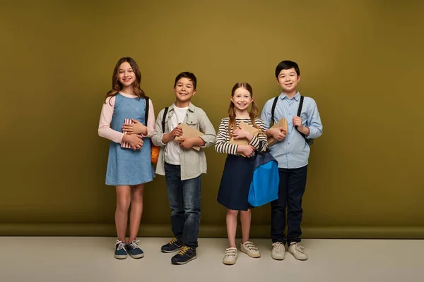 Повна довжина веселих багатоетнічних дев'ятнадцяти дітей у повсякденному одязі, що тримає рюкзаки та книги на фоні хакі, концепція щасливого дитячого дня — стокове фото