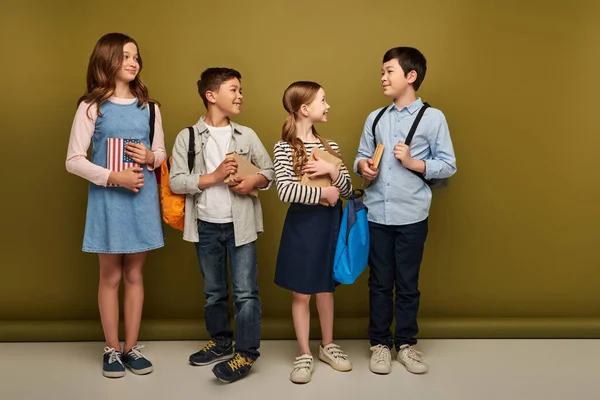 Lächelnde Vorschulkinder, die auf khakifarbenem Hintergrund eine asiatische Freundin mit Rucksack und Buch betrachten, internationales Konzept zum Kinderschutztag — Stockfoto
