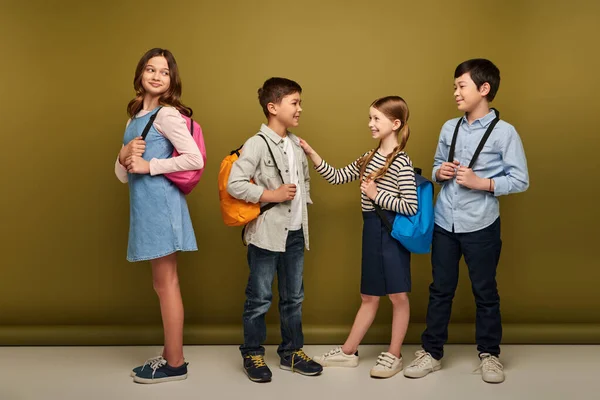 Full length of smiling interracial kids with backpacks looking at friends talking during international child protection day celebration on khaki background — Stock Photo