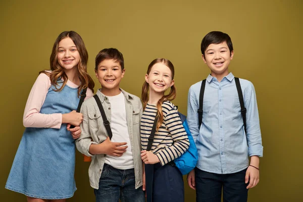 Усміхнені багатоетнічні діти з рюкзаками в повсякденному одязі, дивлячись на камеру, стоячи разом під час святкування Дня захисту дітей на фоні хакі — стокове фото