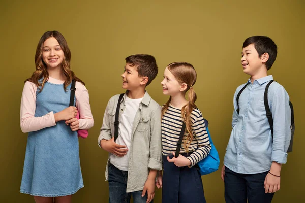Positive Preteen-Kids in Freizeitkleidung betrachten einen Freund im Kleid, der mit Rucksack bei der Feier zum Kinderschutztag auf khakifarbenem Hintergrund steht — Stockfoto