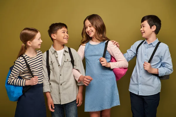 Smiling multiethnic and preteen friends with backpacks talking with each other during child protection day celebration while standing on khaki background — Stock Photo
