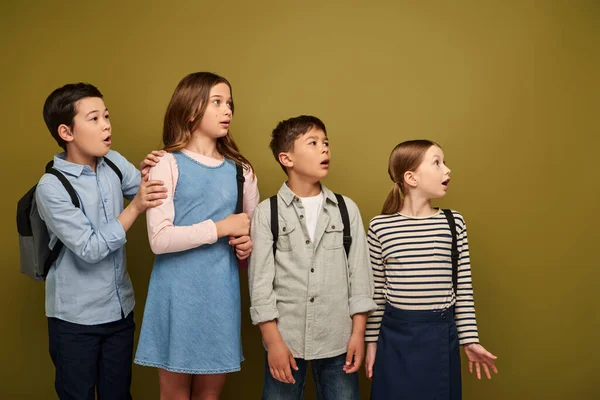 Shocked multiethnic preteen schoolkids in casual clothes with backpacks looking away and opening mouth during child protection day celebration on khaki background — Stock Photo