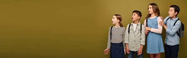 Surprised multicultural schoolkids with backpacks looking away during international child protection day celebration on khaki background with copy space, banner — Stock Photo