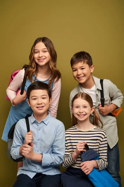 Усміхнені багатоетнічні діти в повсякденному одязі тримають рюкзаки і дивляться на камеру під час святкування дня захисту дітей на фоні хакі — стокове фото