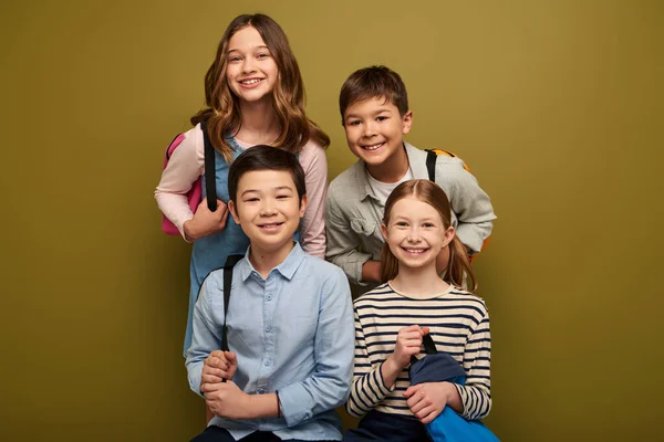 Lächelnde multiethnische Schulkinder mit Rucksäcken, die bei der Feier zum Weltkinderschutztag auf khakifarbenem Hintergrund in die Kamera schauen — Stockfoto