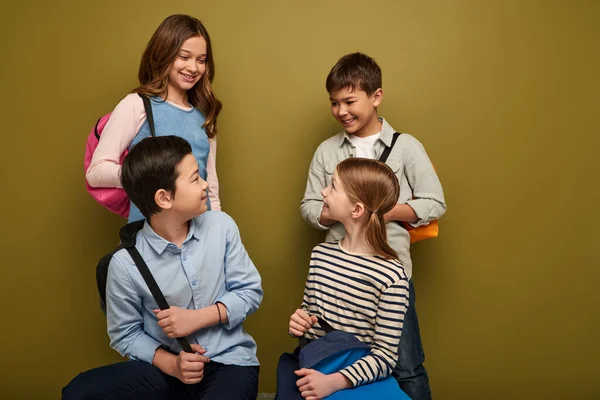 Positive multiethnische Schulkinder unterhalten sich mit ihren Vorschulfreunden, während sie Rucksäcke halten, einander anschauen und den internationalen Kindertag auf khakifarbenem Hintergrund feiern — Stockfoto