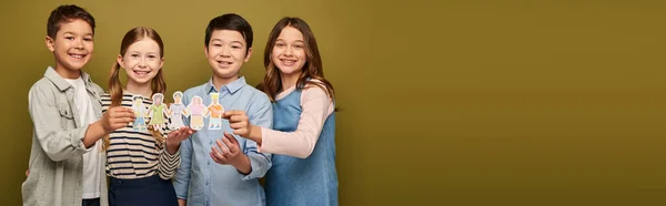 Happy interracial preteen friends in casual clothes holding drawn paper craft characters and looking at camera during child protection day celebration on khaki background with copy space, banner — Stock Photo