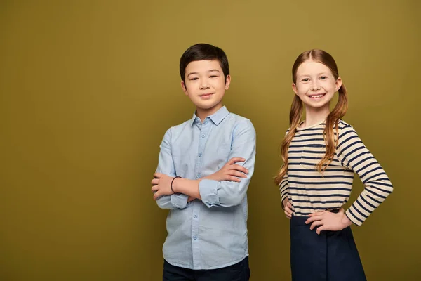 Smiling redhead girl holding hands on hips and looking at camera near asian friend crossing arms during child protection day celebration on khaki background — Stock Photo