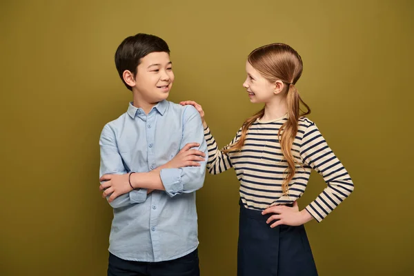 Smiling redhead preteen girl hugging and looking at asian friend folding hands during international child protection day celebration on khaki background — Stock Photo