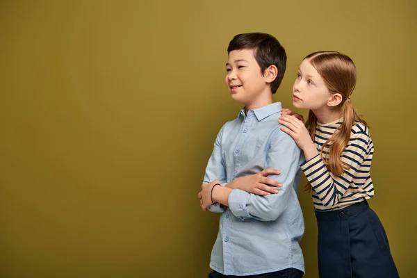 Redhead girl in striped blouse hugging smiling asian friend crossing and and looking away during child protection day celebration on khaki background with copy space — Stock Photo