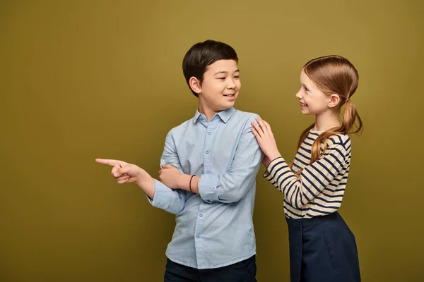 Lächelndes rothaariges Mädchen in gestreifter Bluse umarmt eine asiatische Freundin, die mit dem Finger wegzeigt, während sie etwas während der Kinderschutztagsfeier auf khakifarbenem Hintergrund zeigt — Stockfoto