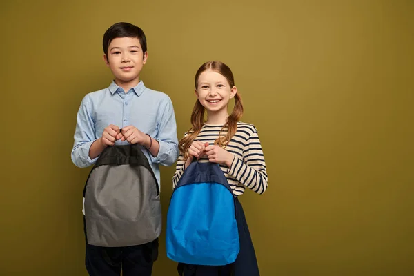Positive redhead preteen girl holding backpack near asian friend in shirt and looking at camera together during child protection day celebration on khaki background — Stock Photo