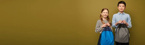 Sonriente chica pelirroja sosteniendo la mochila cerca de preadolescente amigo asiático durante la celebración del día de protección infantil en fondo caqui con espacio de copia, pancarta - foto de stock
