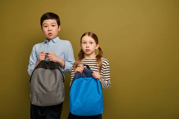 Scared multiethnic preteen kids in casual clothes holding backpacks and looking at camera together during international child protection day celebration on khaki background — Stock Photo
