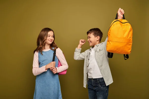 Захоплений багаторасовий хлопчик тримає рюкзак і показує так жест біля друга в одязі, що стоїть і посміхається під час святкування дня захисту дітей на фоні хакі — стокове фото