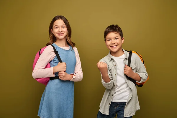 Excited multiracial boy with backpack showing yes gesture and looking at camera near friend in dress during child protection day celebration on khaki background — Stock Photo
