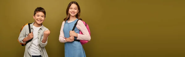 Carefree multiracial boy with backpack showing yes gesture at camera near friend during child protection day celebration on khaki background with copy space, banner — Stock Photo