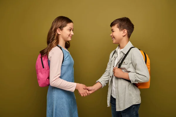 Smiling multiethnic schoolkids with backpacks shaking hands and looking at each other during international child protection day celebration on khaki background — Stock Photo