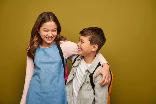 Carefree preteen and multiethnic kids in casual clothes with backpacks hugging each other and smiling during global child protection day on khaki background — Stock Photo