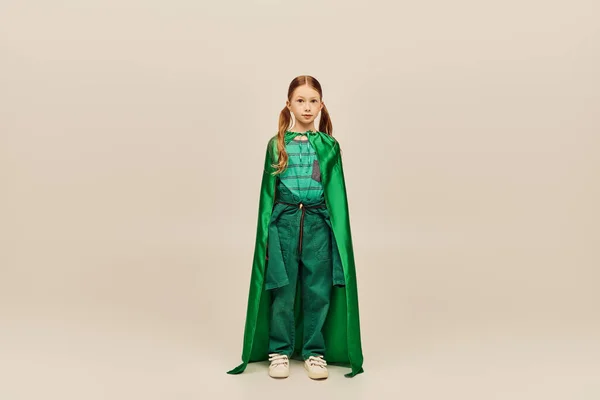 Руда дівчина в зеленому костюмі супергероя і накидка дивиться на камеру, стоячи на сірому фоні під час святкування Всесвітнього дня захисту дітей — стокове фото