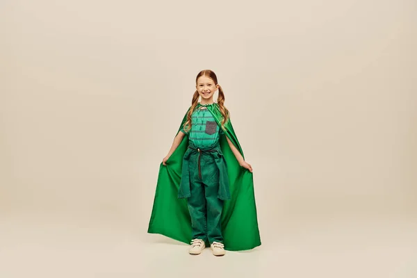 Cheerful redhead kid in green superhero costume holding cape and looking at camera while standing on grey background in studio during international children`s day celebration — Stock Photo