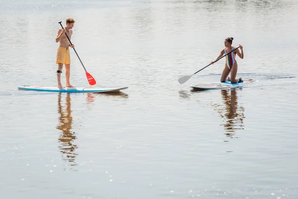 Sportive african american woman in colorful swimsuit and young redhead man in swim shorts spending summer weekend while sailing on sup boards together — Stock Photo