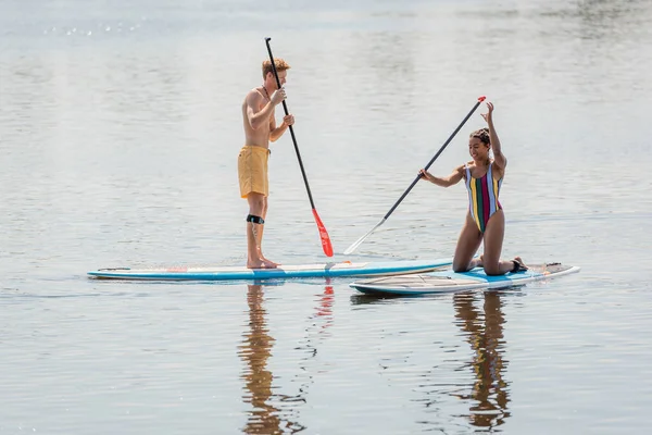 Femme afro-américaine gaie en maillot de bain coloré agenouillé sur la planche à soup près de rousse homme naviguant avec pagaie tout en passant du temps sur la rivière en été — Photo de stock