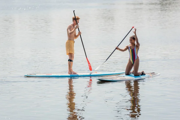 Alegre africana americana mujer en colorido traje de baño arrodillado en sup board cerca pelirrojo hombre vela con remo mientras pasar tiempo en el río en verano — Stock Photo