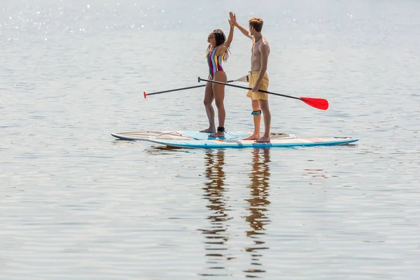 Positive and sportive redhead man and african american woman in colorful swimwear holding paddles and giving high five while sailing on sup boards on lake in summer — Stock Photo