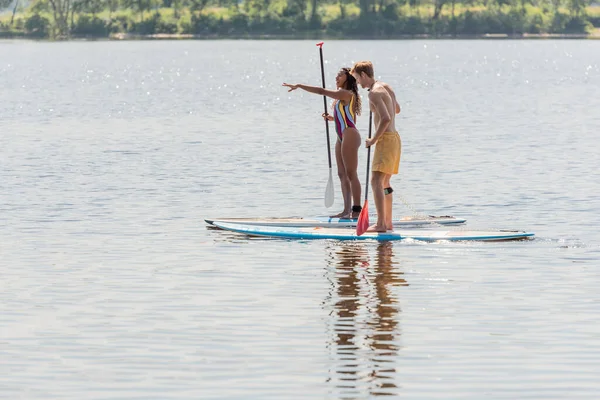 Vista lateral de la mujer afroamericana en traje de baño colorido apuntando con la mano cerca de hombre joven deportivo mientras navega en tablas de sup durante el fin de semana en el lago - foto de stock