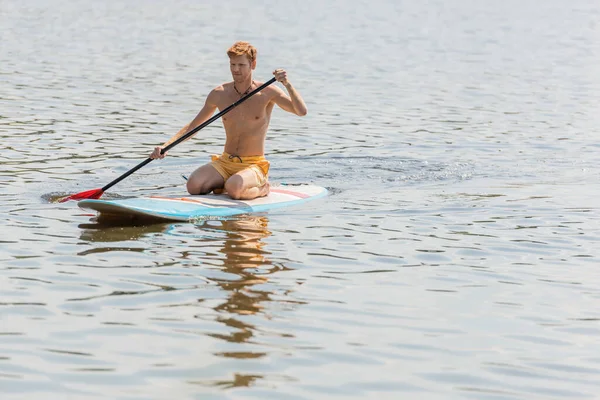 Young and sportive redhead man in yellow swim shorts spending summer weekend on river while sitting on sup board and sailing with paddle during water recreation — Stock Photo