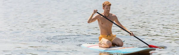 Young and active redhead man in yellow swim shorts spending summer weekend on river while sitting on sup board and sailing with paddle during water recreation, banner — Stock Photo