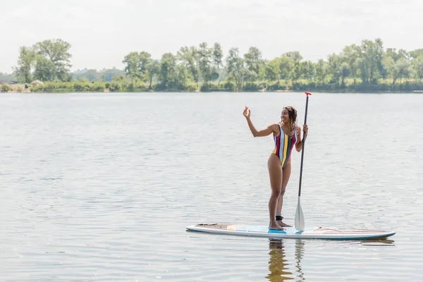 Full length of carefree african american woman in striped swimsuit standing with paddle on sup board and waving hand while looking away on lake on summer day with picturesque riverside on background — Stock Photo