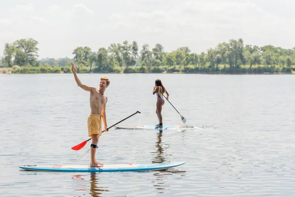 Веселий рудий чоловік дивиться в сторону і вказує рукою біля спортсменки афроамериканської жінки в барвистому купальнику, що плаває на борту в літній день з мальовничим берегом річки на фоні — стокове фото