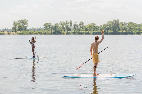 Active interracial couple in colorful swimwear looking at each other and waving hands while sailing on sup boards on picturesque lake with green shore — Stock Photo
