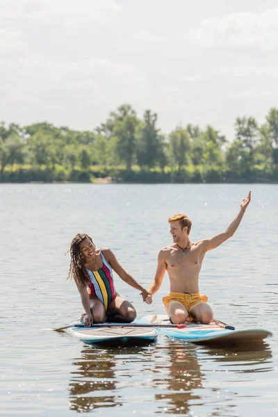 Homme rousse insouciant tenant la main avec femme afro-américaine gaie et pointant du doigt tout en étant assis sur des planches à soup sur le lac avec une banque pittoresque verte — Photo de stock