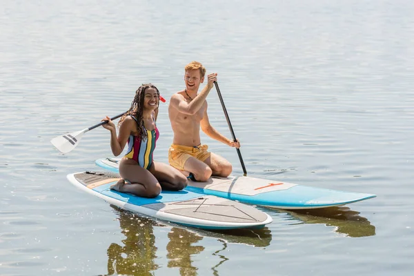 Mujer afroamericana llena de alegría en traje de baño a rayas y hombre pelirrojo feliz en pantalones cortos de baño sentado en tablas sup mientras pasa tiempo en el lago en el fin de semana de verano - foto de stock