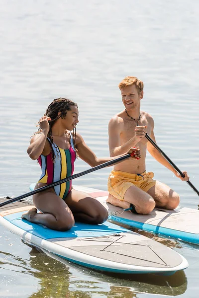 Happy redhead man looking at enchanting african american woman in colorful swimsuit sailing on sup board while spending time together on lake in summer — Stock Photo