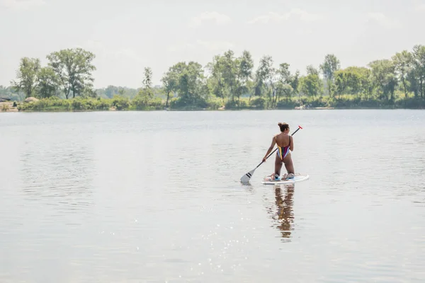 Vue arrière de la femme afro-américaine active en maillot de bain rayé se reposant sur la rivière en naviguant sur un tableau près de la rivière avec des arbres verts, une activité de plein air, des vibrations estivales — Photo de stock