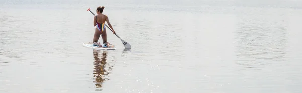 Rückansicht einer afrikanisch-amerikanischen Frau in farbenfrohem und gestreiftem Badeanzug, die Zeit auf dem See verbringt, indem sie auf einem Sup Board mit Paddel, Banner, Outdoor-Aktivitäten, Sommerstimmung segelt — Stockfoto