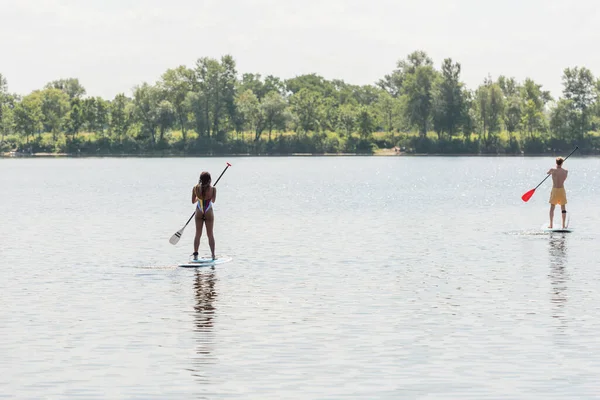 Задний вид спортивного мужчины и африканской американки в красочных купальниках, плавающих на досках с веслами на озере с зеленым живописным берегом, активностью на открытом воздухе, летними вибрациями — стоковое фото