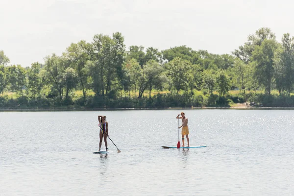 Full length of sportive african american woman with young friend sailing on sup boards with paddles along riverside with green trees during summer weekend — Stock Photo