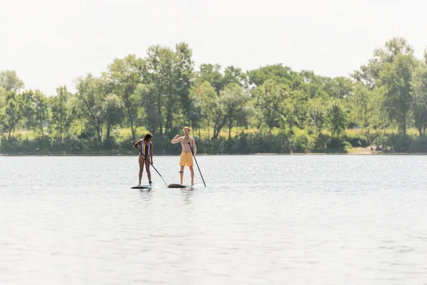 Full length of sportive and active interracial couple spending time on lake while sailing on sup boards with paddles along shore with green summer trees — Stock Photo