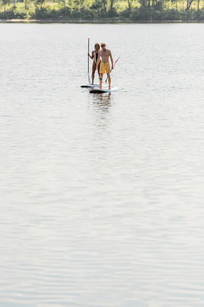 Активная многонациональная пара в красочных купальниках, плывущая на досках с веслами на спокойной речной воде возле зеленого берега во время летнего водного отдыха — стоковое фото