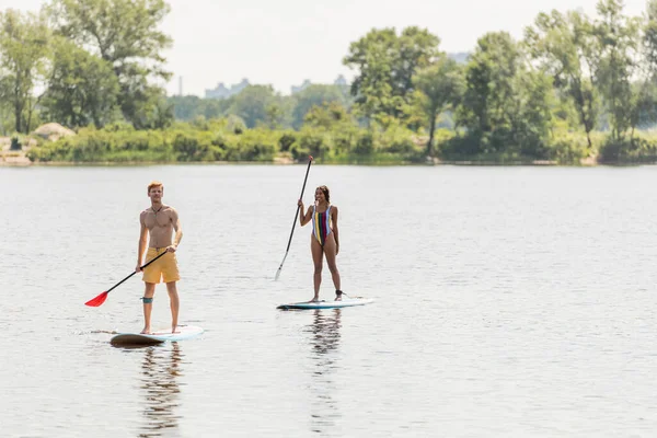 Redhead man in swim shorts and african american woman in colorful swimsuit holding paddles while sailing on sup boards along green shore on summer day — Stock Photo