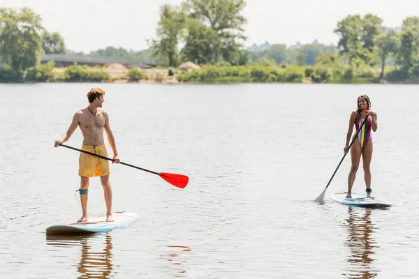 Full length of interracial sportive couple holding paddles and smiling at each other while sailing on sup boards on river during water recreation on summer weekend — Stock Photo
