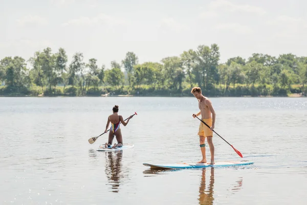 Young and sportive redhead man standing on sup board with paddle near african american woman in colorful swimsuit sailing on picturesque lake with green shore — Stock Photo