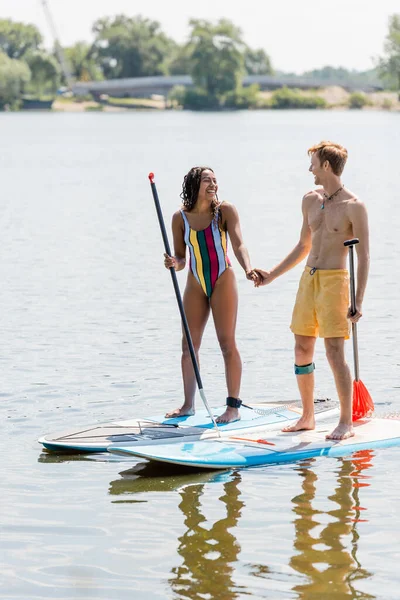 Active redhead man and cheerful african american woman in striped swimsuit holding hands and smiling at each other while standing on sup boards on lake in summer — Stock Photo